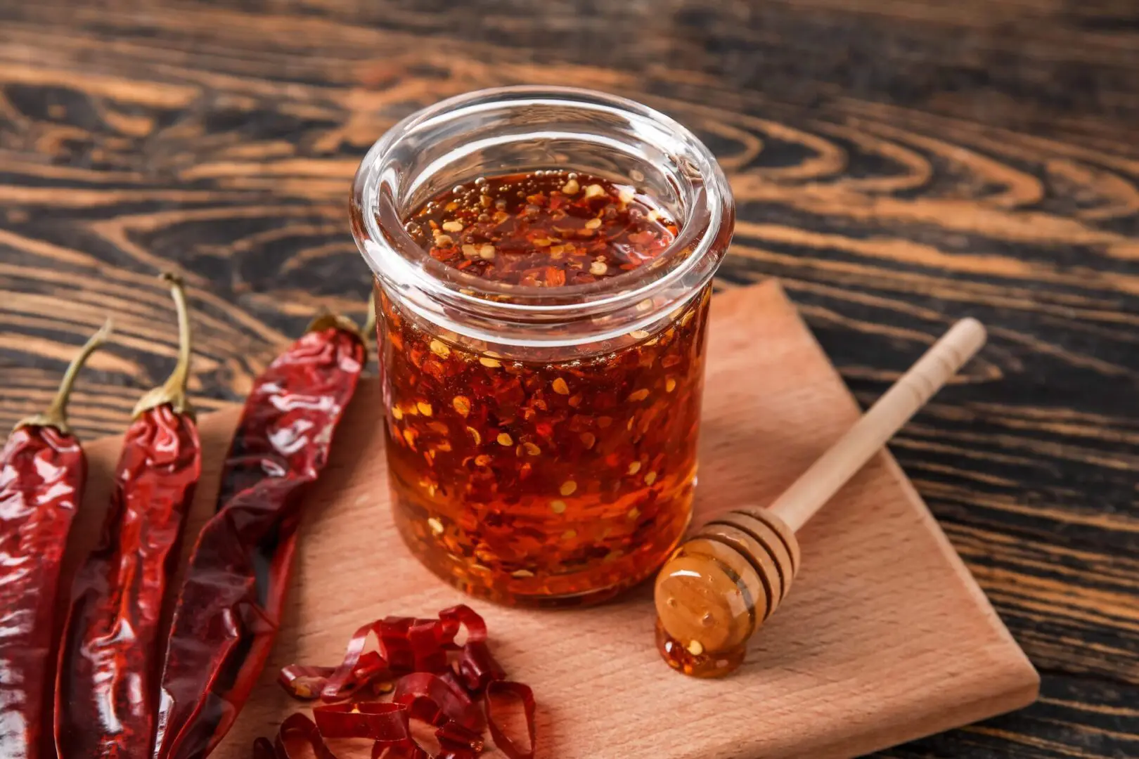 A clear glass jar filled with rich, golden-amber homemade wildflower hot honey infused with red chili flakes, sitting on a rustic wooden kitchen surface alongside fresh red chili peppers.