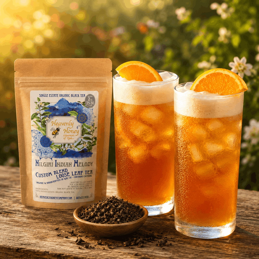Two tall, frosted glasses of ice-shaken Ojai Wildflower Nilgiri Tea topped with a light foam layer and garnished with fresh orange slices, sitting on a rustic outdoor table under soft golden hour sunlight.