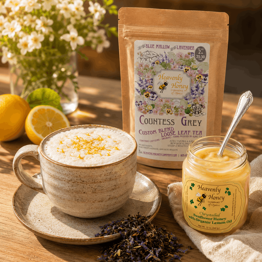 A wide ceramic mug filled with a frothy Ojai Countess Creamed Citrus Latte topped with a thick foam layer and a pinch of lemon zest, sitting on a sunlit wood table next to an open glass jar of velvety creamed lemon honey with a silver spoon.