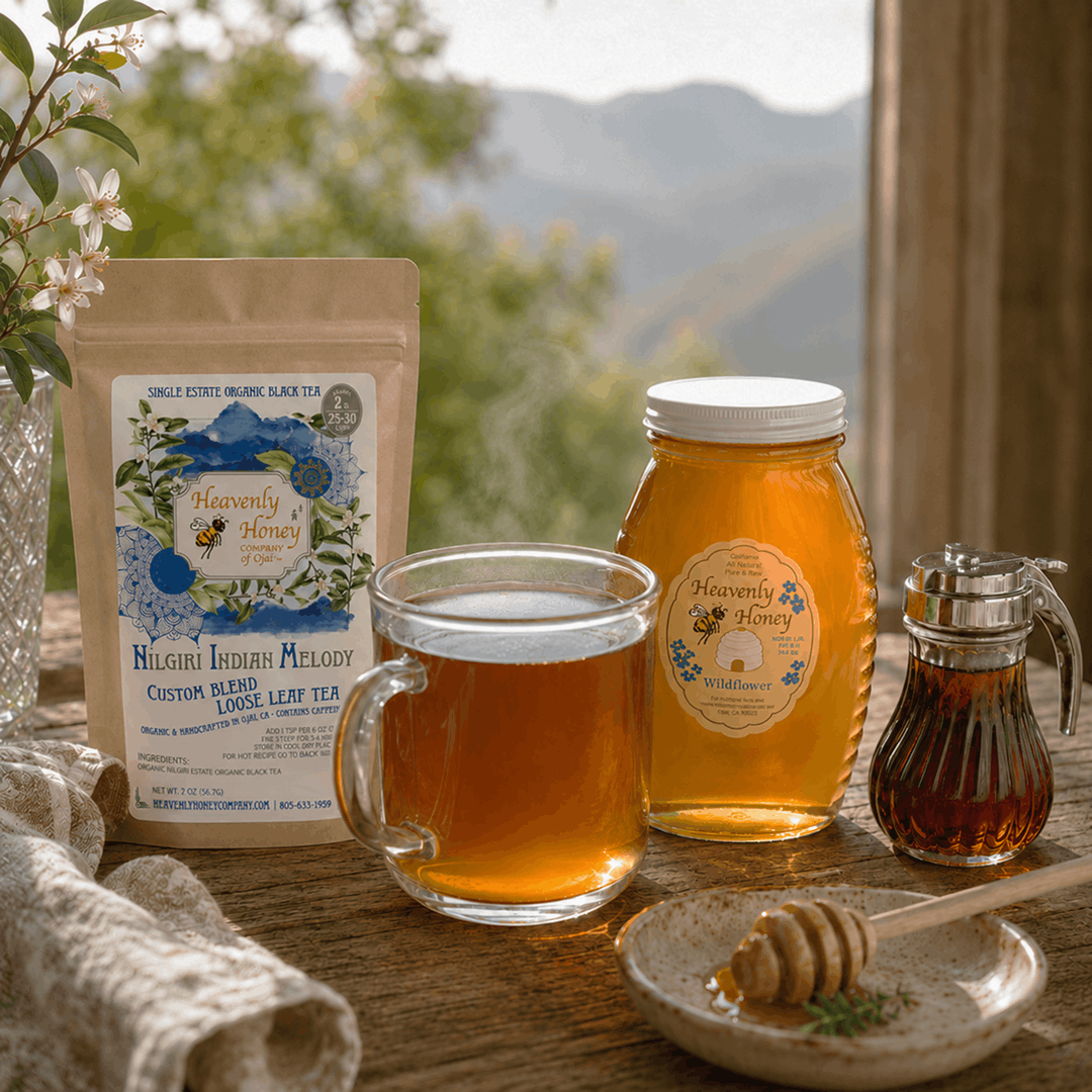 A clear glass mug filled with amber-colored Ojai Ethical Nilgiri Blossom Brew tea, casting soft shadows on a sunlit wooden table. A rustic jar of Wildflower honey and a small pourer of maple syrup sit neatly next to the mug under warm afternoon light.