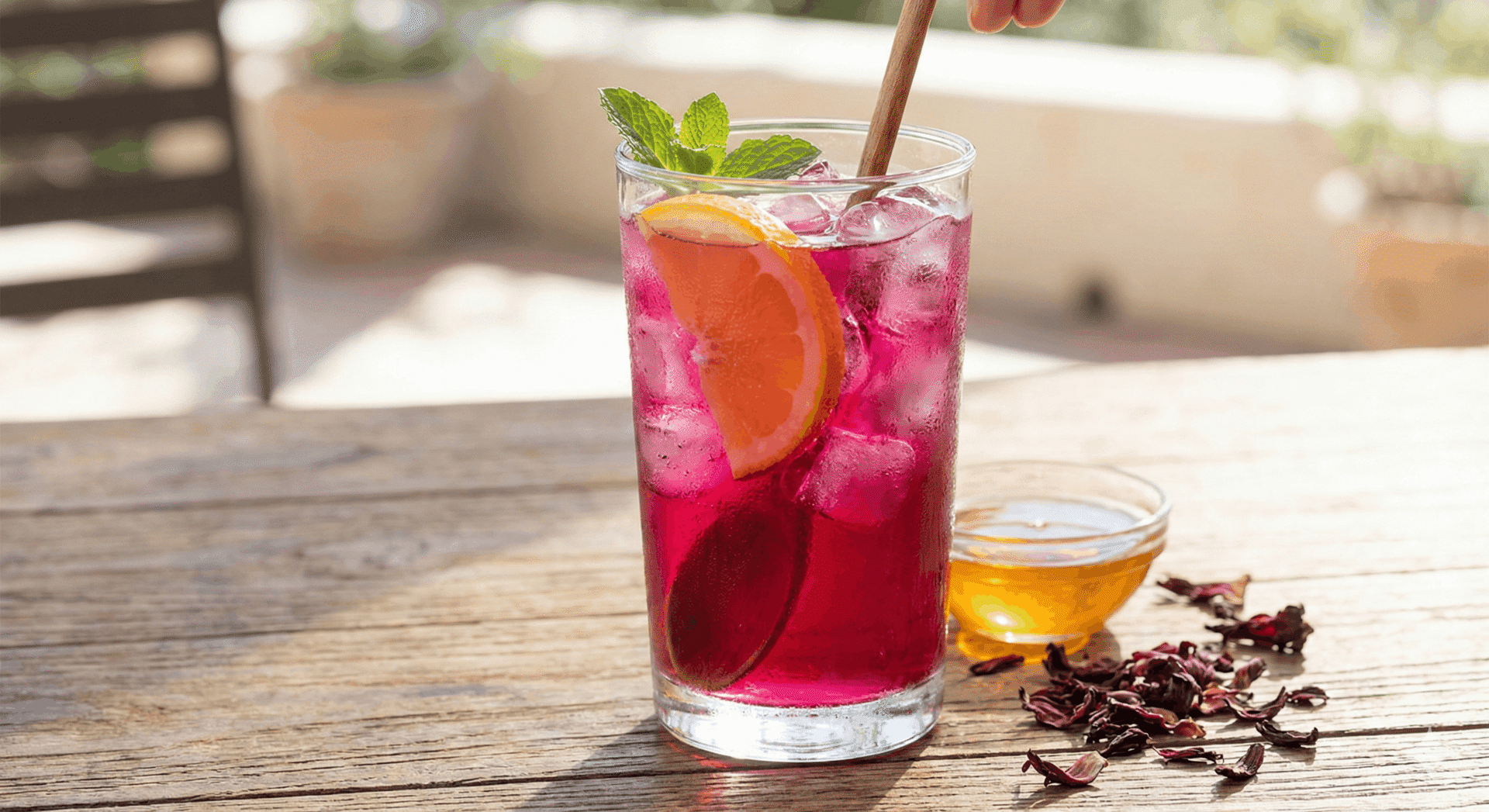 A tall glass of vibrant pink Hibiscus & Orange Blossom Iced Cooler with ice cubes, an orange wedge, and fresh mint.